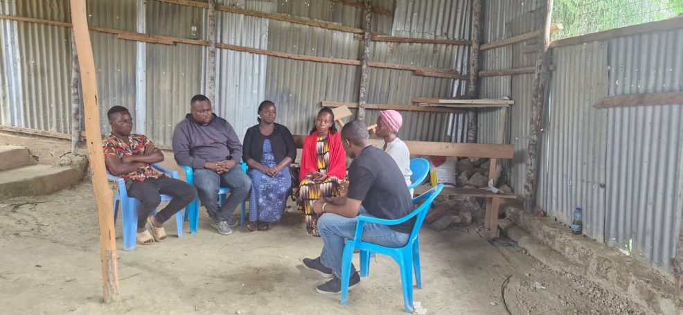 Eine Gruppe von sechs Personen sitzt in einem einfachen Raum auf blauen Stühlen.